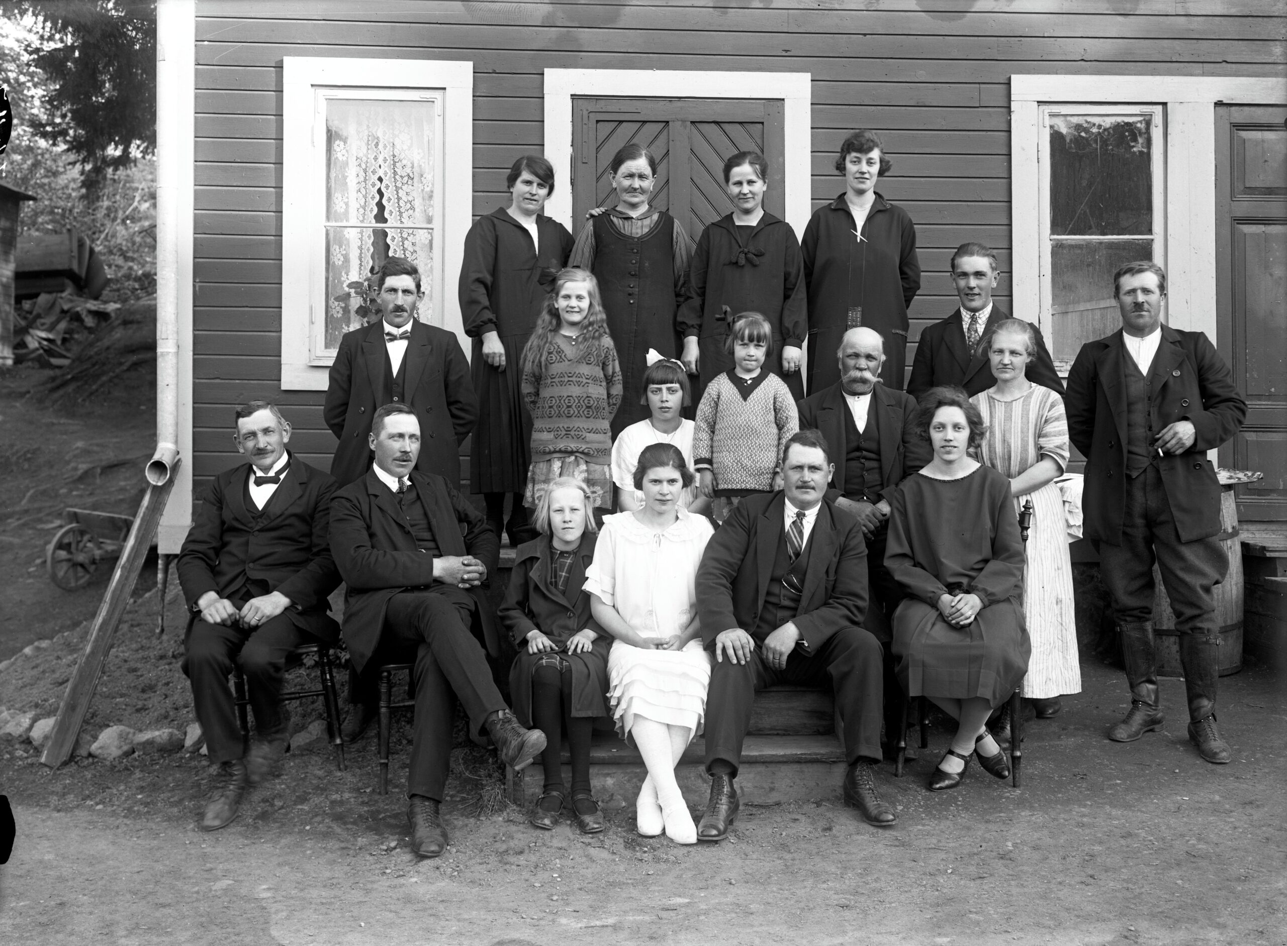 Svartvitt familjefoto där 18 personer står uppställda framför ett trähus.