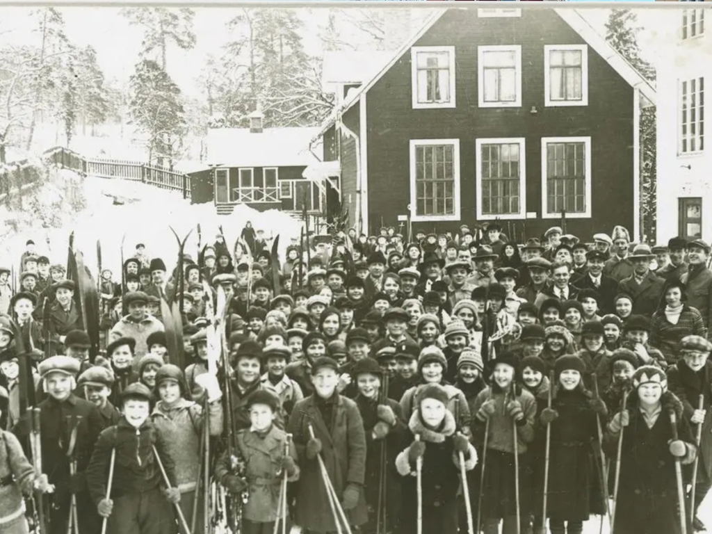 Ett hundratal barn står uppställda med vinterkläder och skidor framför skolhuset.