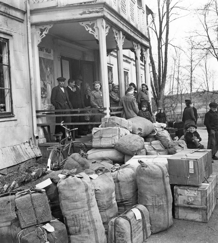 Män, kvinnor och barn står på en veranda. Massor av baggage runt omkring.