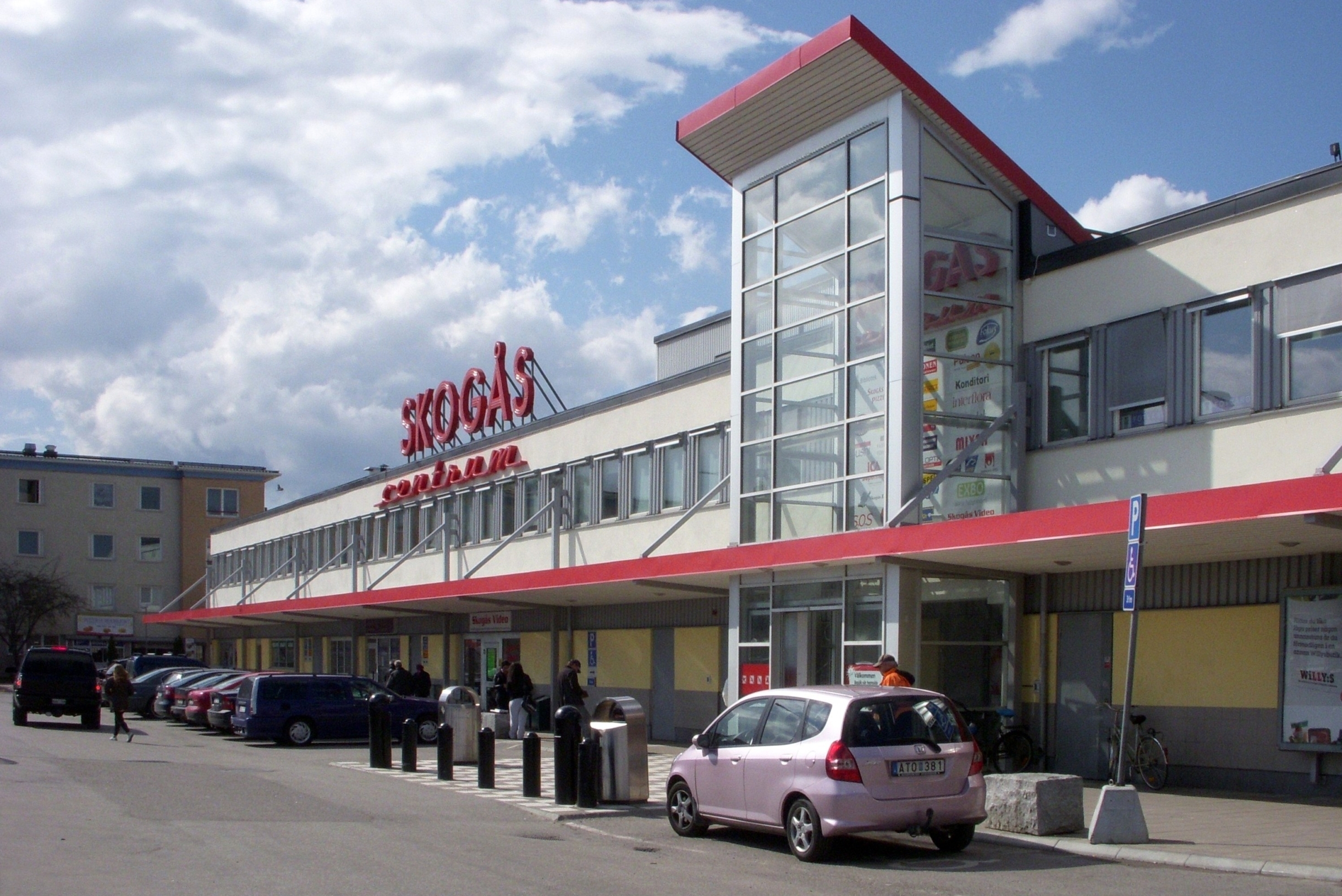 Färgfoto av centrumbyggnaden med skylten Skogås centrum. Röda detaljer mot blå himmel.