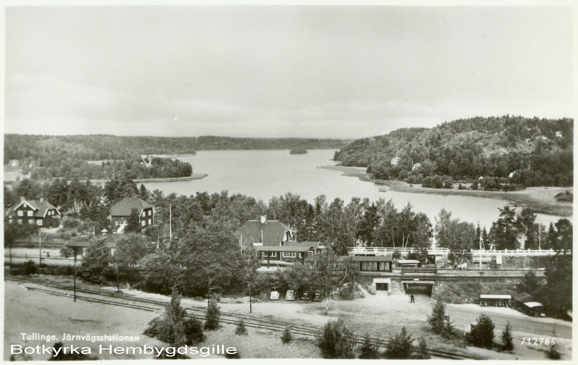 Järnvägsspåren och perrongen i förgrunden. Intill syns stationshus och andra hus och vyn är vidsträckt över Tullingesjön i bakgrunden.