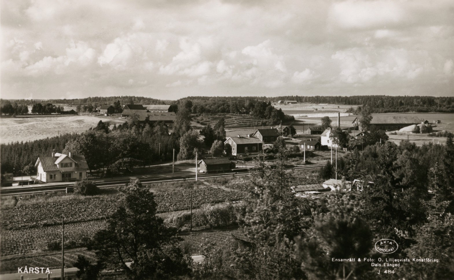Svartvit bild. Enstaka hus och jordbrukslandskap i bakgrunden.