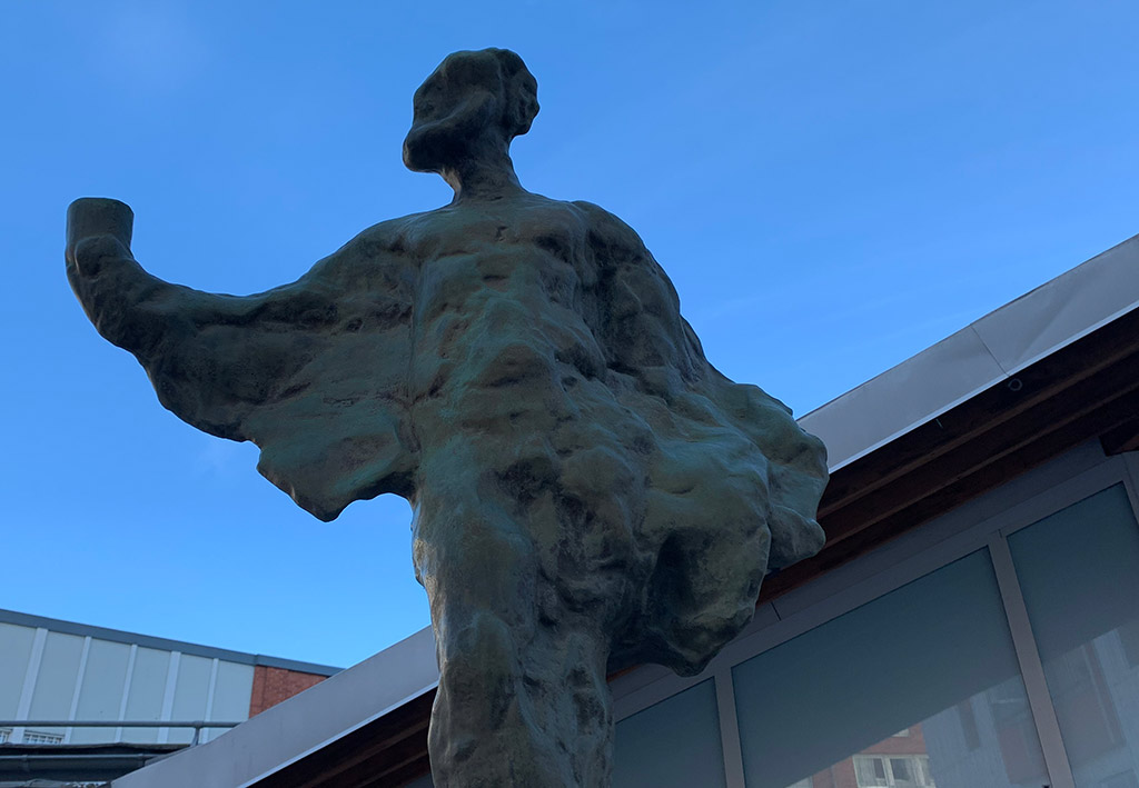 Skulptur av en människa i brons som höjer en bägare mot en blå himmel. Abstrakta former.