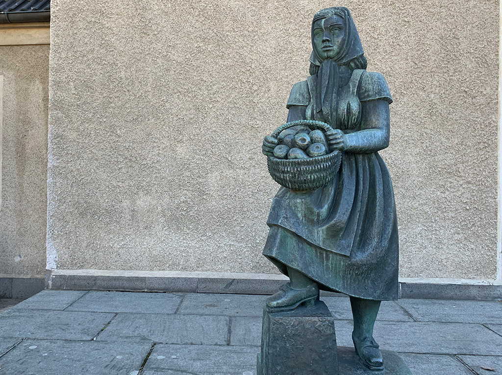 Skulptur av en kvinna med sjalett och klänning med en fruktkorg i händerna. Gjord av brons och står mot en beige putsad vägg.
