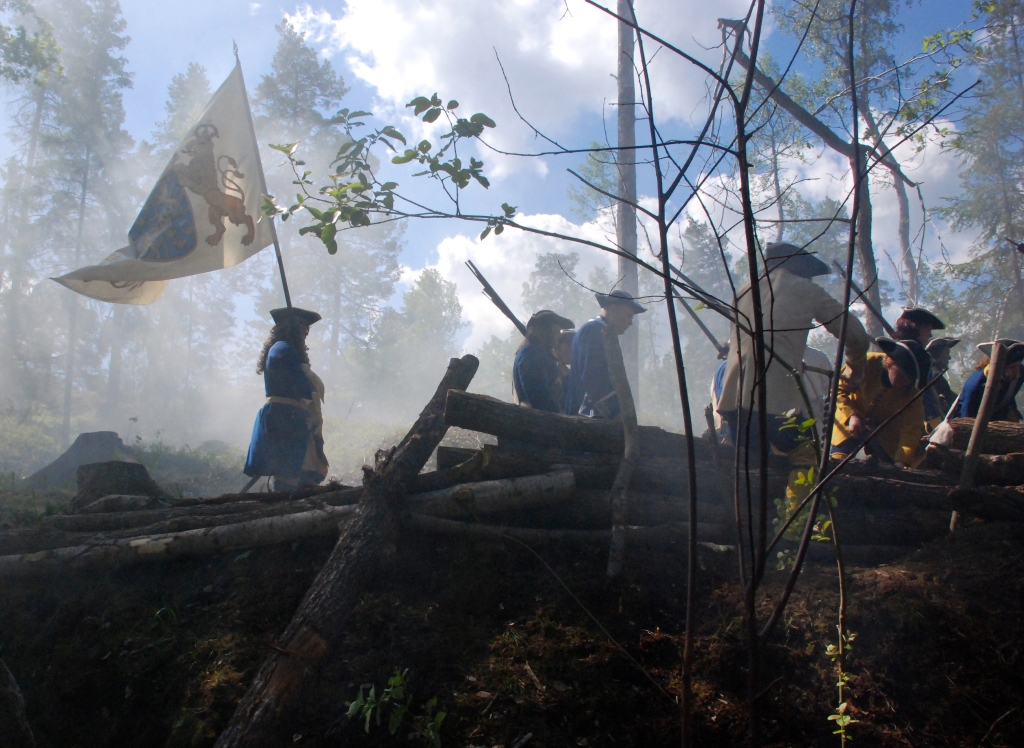 Soldater i blå uniformer med gevär och en flagga. Stockar på marken och rök i luften.