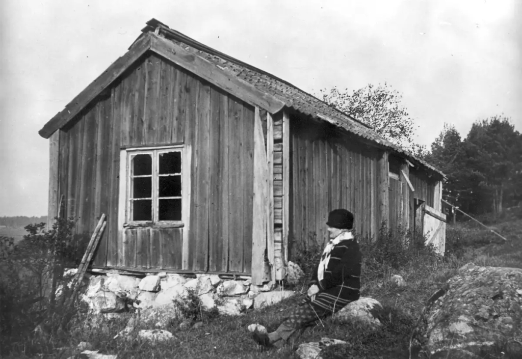 Svartvitt fotografi. Torp med kvinna sittande utanför.