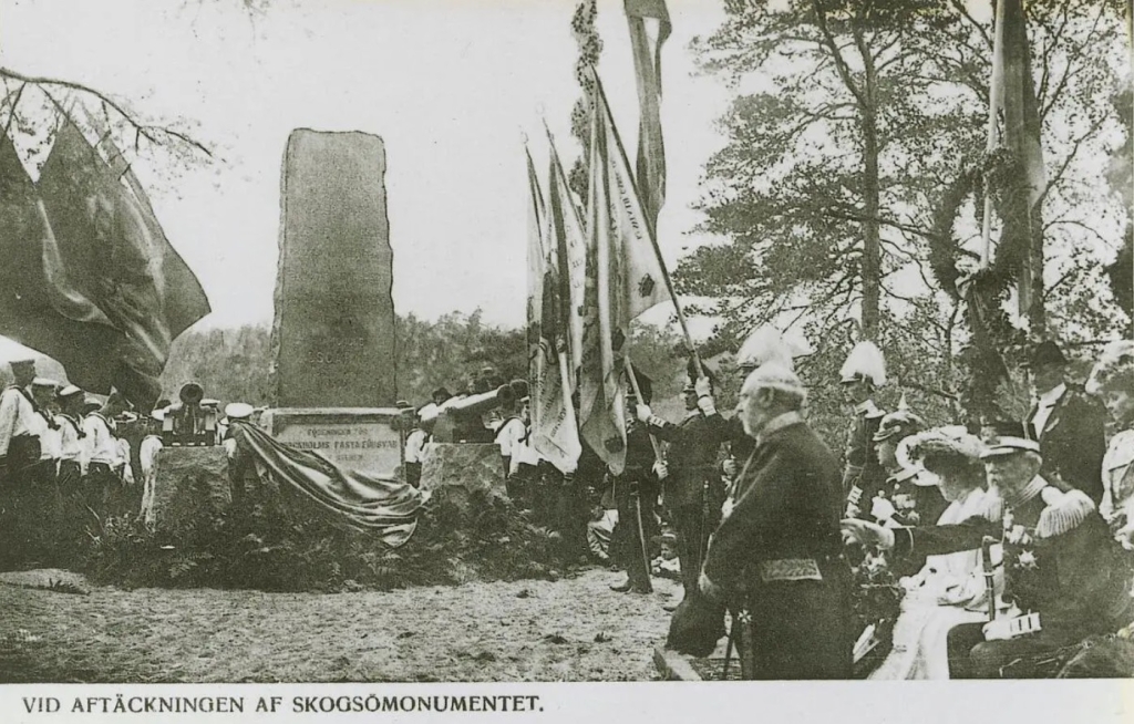 Täckningen faller från stenmonumentet. Militärer med flaggor och kransar. Kungaparet sittande.