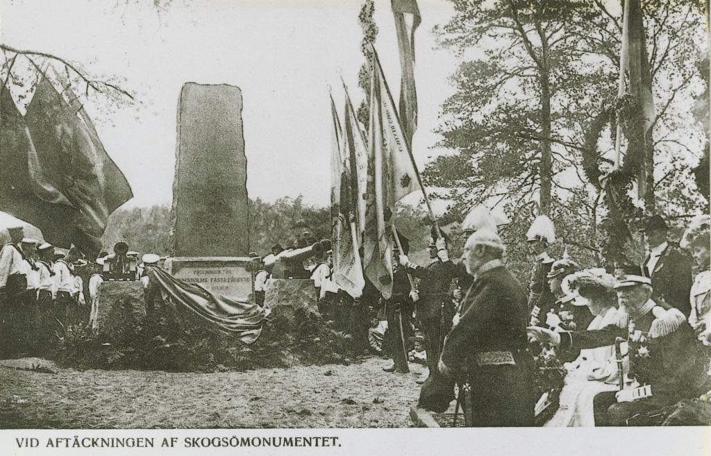 Täckningen faller från stenmonumentet. Militärer med flaggor och kransar. Kungaparet sittande.