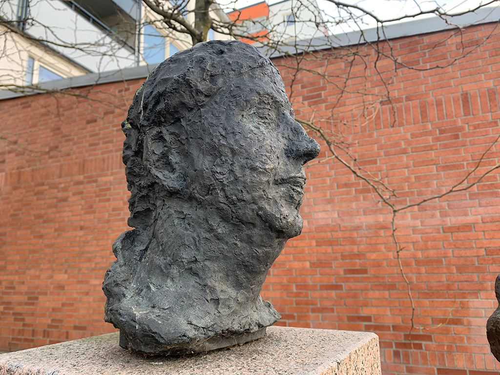 Skulptur av kvinnohuvud i brons på en sockel. I bakgrunden en röd tegelvägg.