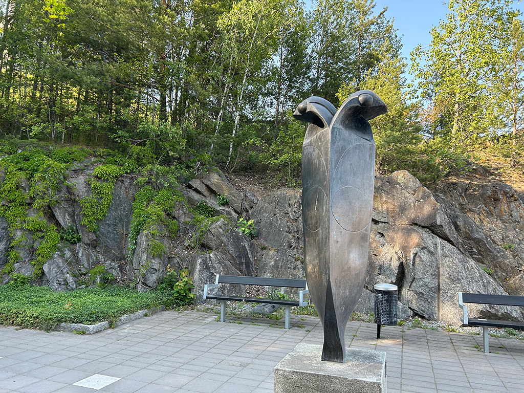 Slät hög skulptur i mörk brons med två huvuden och utstickande runda ögon framför ett berg.