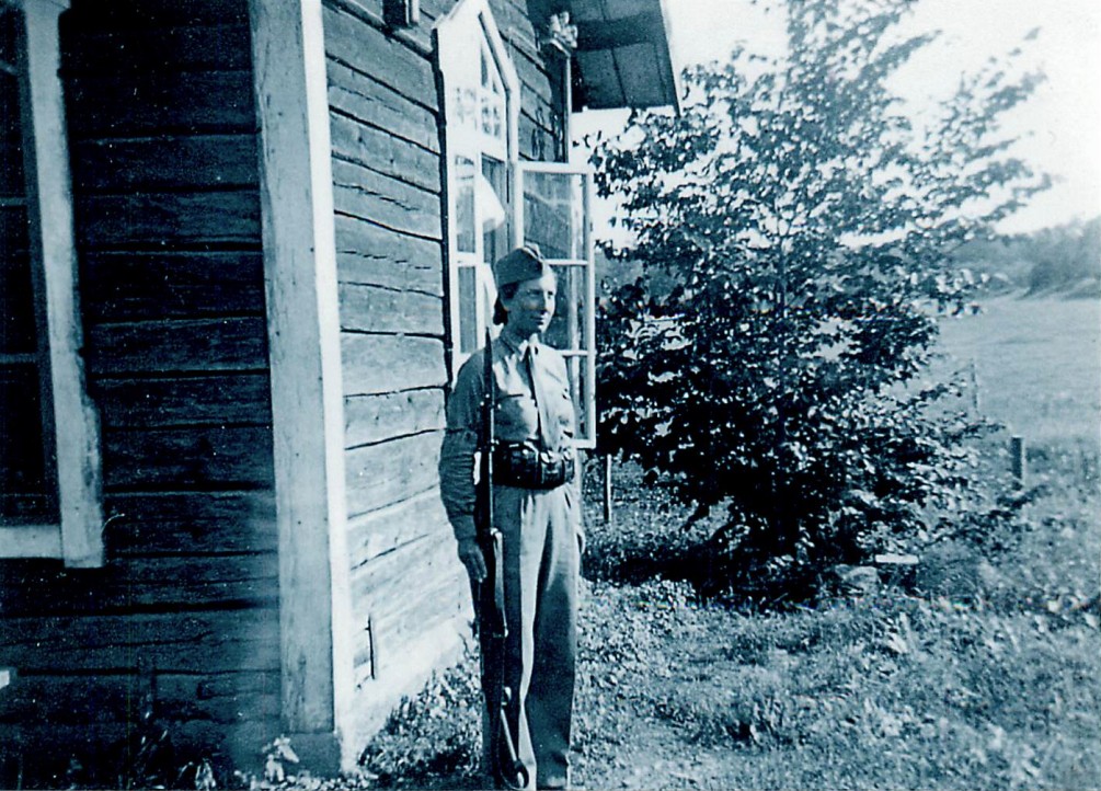 En kvinna i uniform står i givakt med sitt gevär vid en husknut. Timmrat hus, öppet fönster, lantlig omgivning.