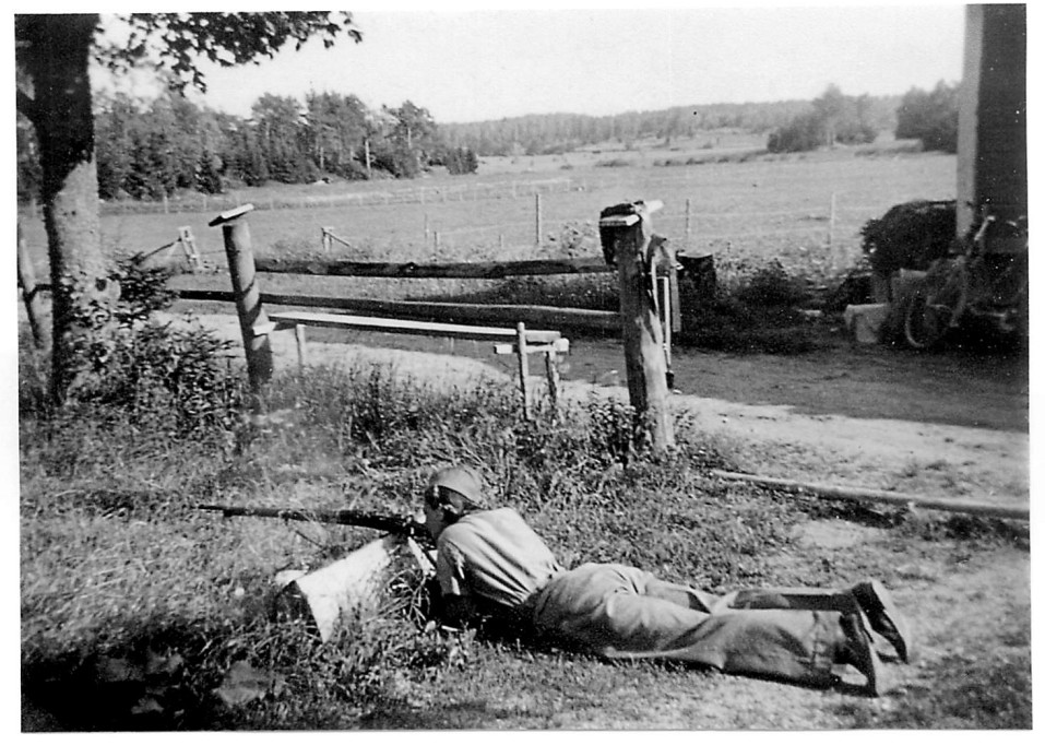 En kvinna ligger på mage på marken och siktar med ett gevär. Jordbrukslandskap i bakgrunden.