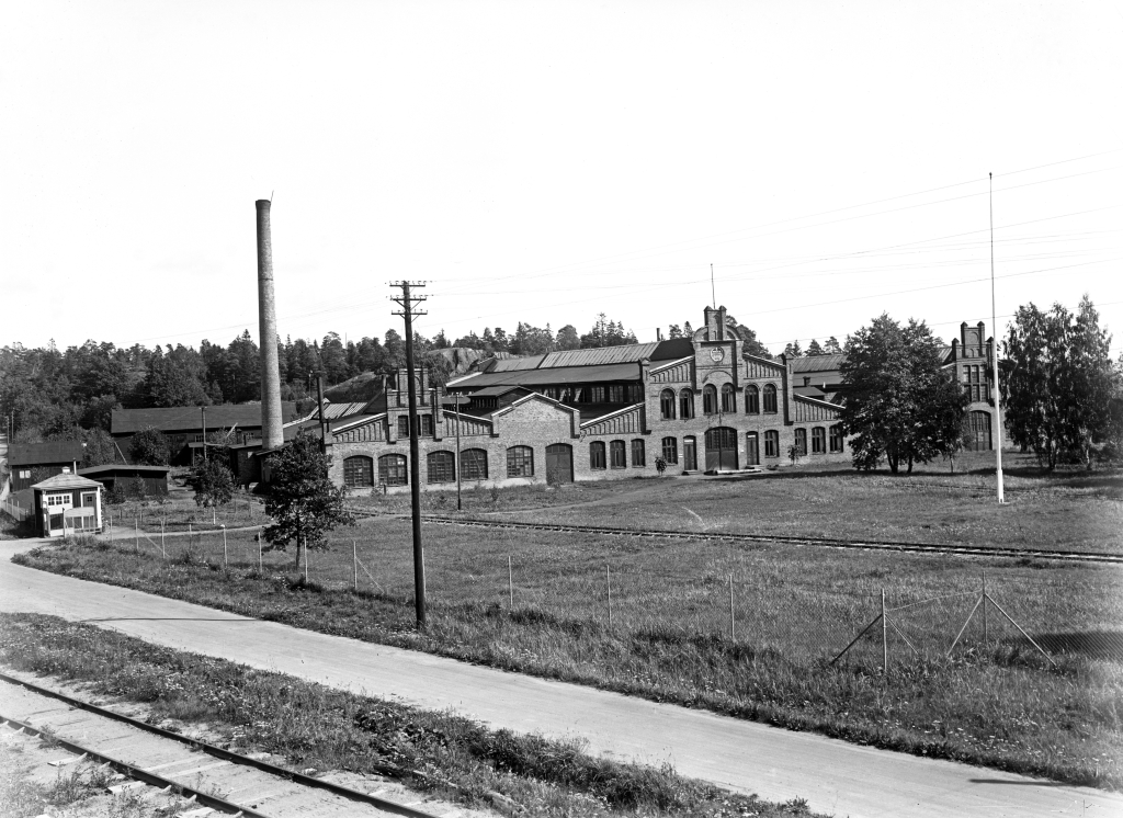 Svartvit bild. Tegelbyggnaden med skorsten. I förgrunden järnväg och stängsel.