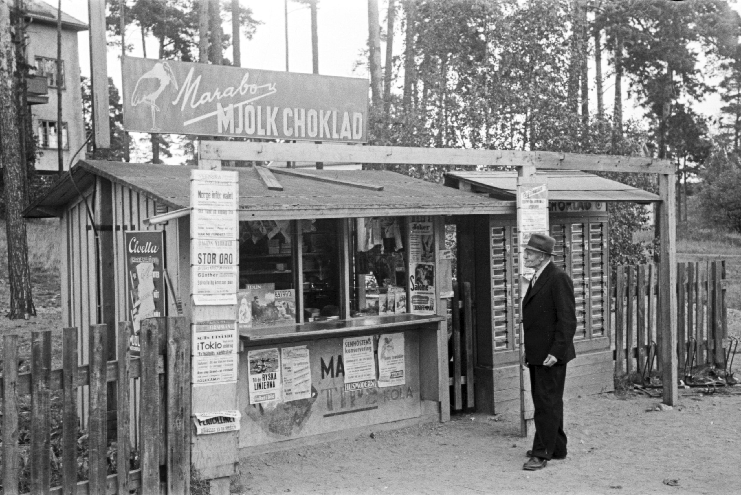 En man i hatt och kostym framför en liten kiosk med tidningslöpsedlar och en stor skylt med texten Marabou Mjölkchoklad.