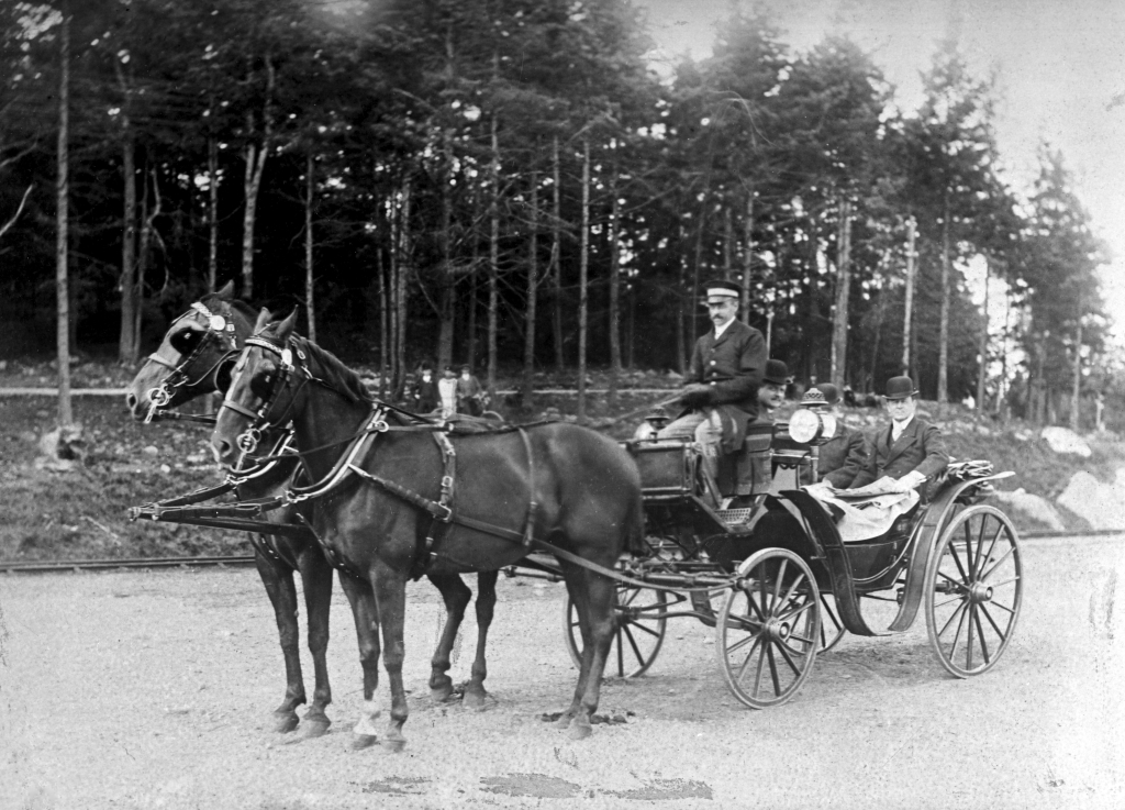 Tre herrar med hatt i en vagn som körs av en kusk och dras av två hästar.