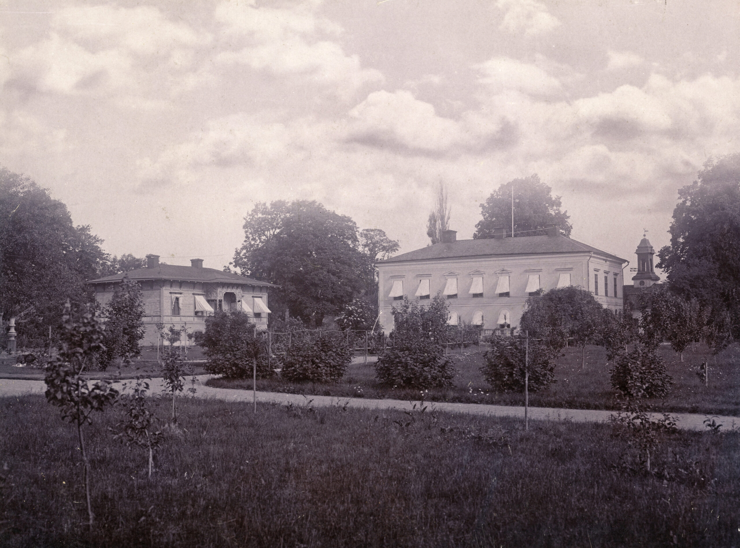 Gårdens huvudbyggnad och en flygelbyggnad. Grönska och nyplanterade träd.