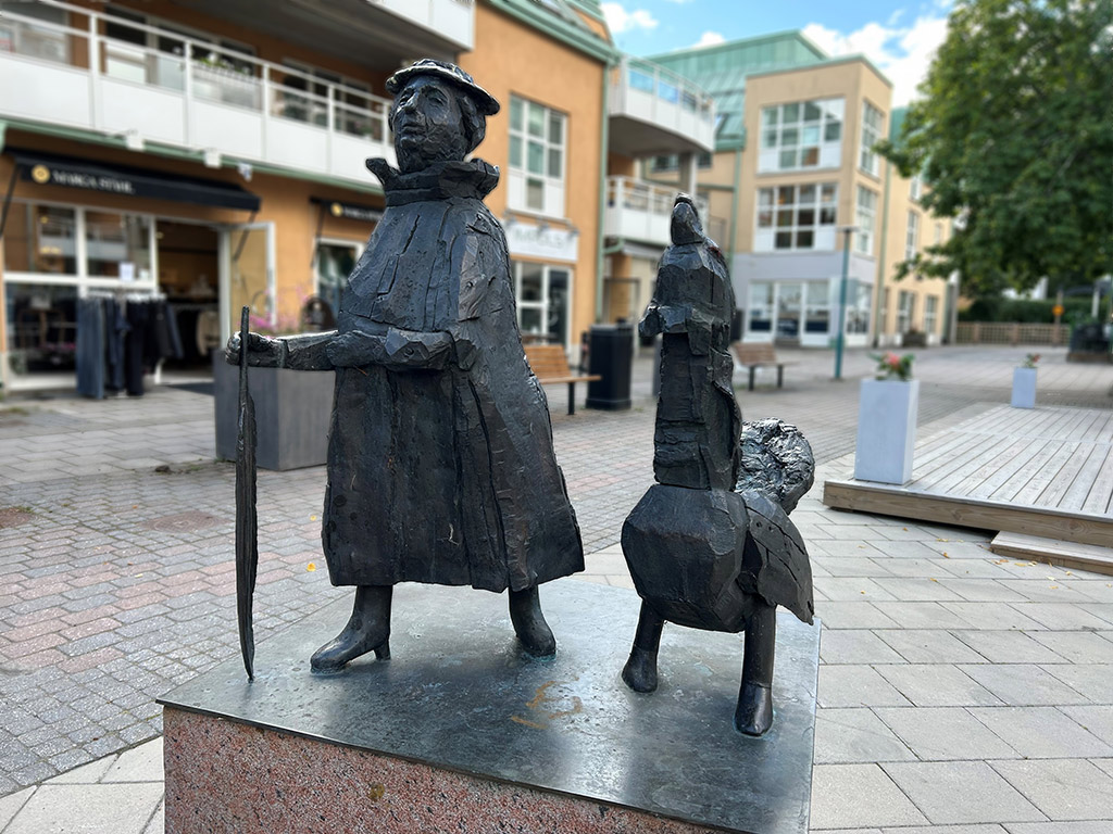 Skulptur av en tant med hatt och kappa och med en fågel bredvid.