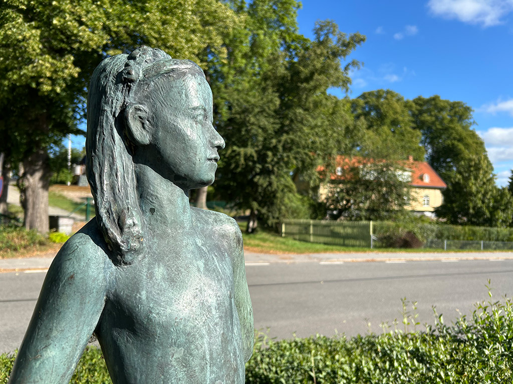 Bronsskulptur av en flicka med långt hår och diadem. Grönskande träd, hus och en väg bakom.