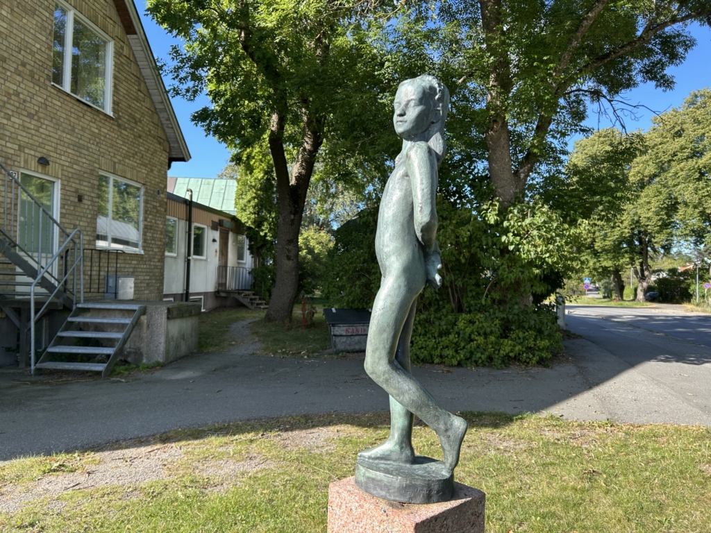 En skulptur av en flicka utanför en gul tegelbyggnad. Grönskande träd bakom.