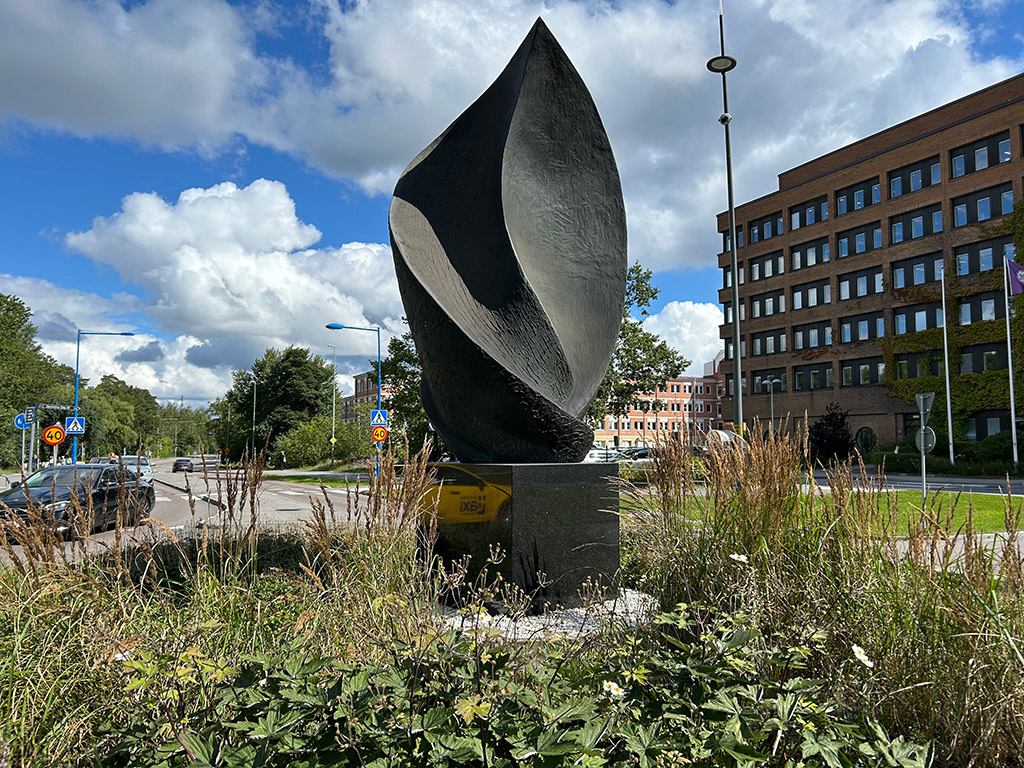 En hög abstrakt bronsskulptur i en rondell med frodig växtlighet. Vägar med bilar och hus runtomkring.