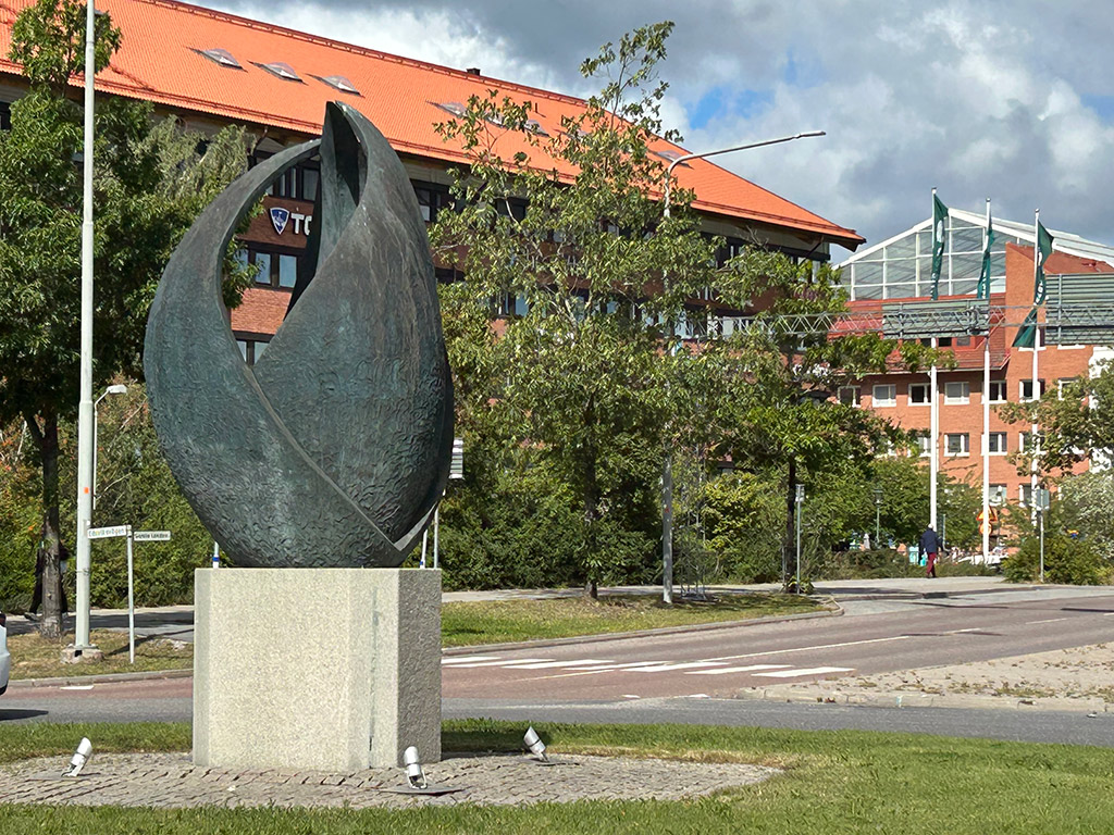Hög bronsskulptur på en vit sockel i en rondell med vägar runtomkring. Hus och grönska i bakgrunden
