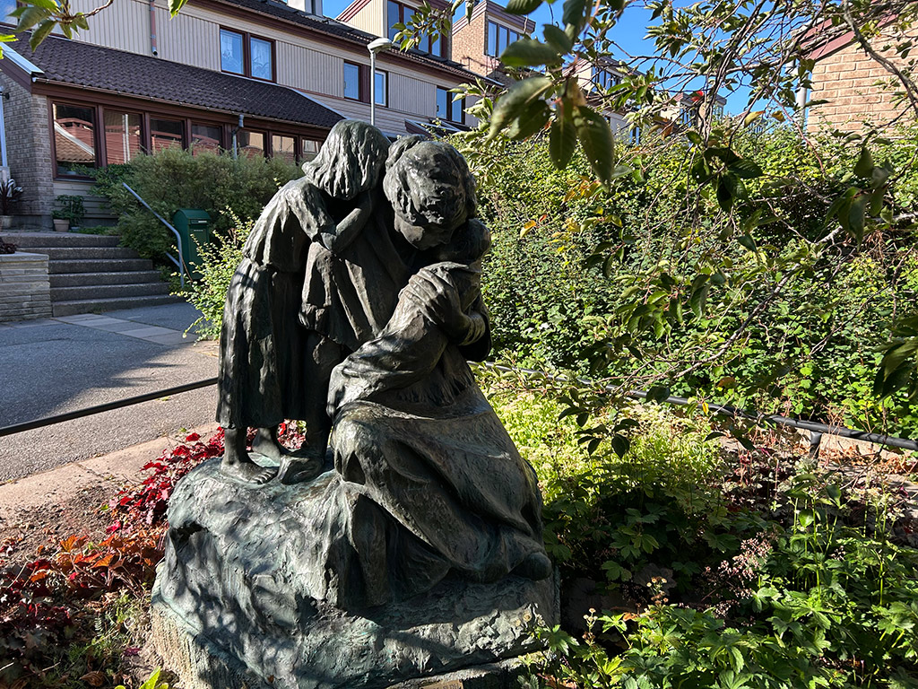 Skulpturgrupp med en kvinna som håller en bebis och en flicka som står bakom kvinnan. I en par med grönska.