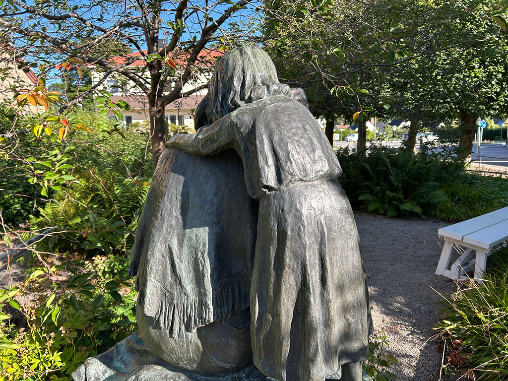 En skulptur av en flicka som håller om en person som sitter ner. I en park med en grusgång och grönska.