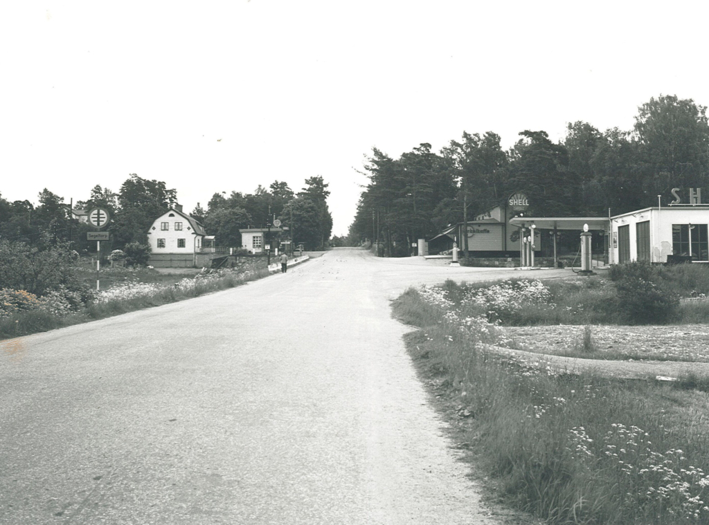 Svartvitt foto. Shellmack, konsum och en villa kantar den asfalterade vägen utan målade streck.