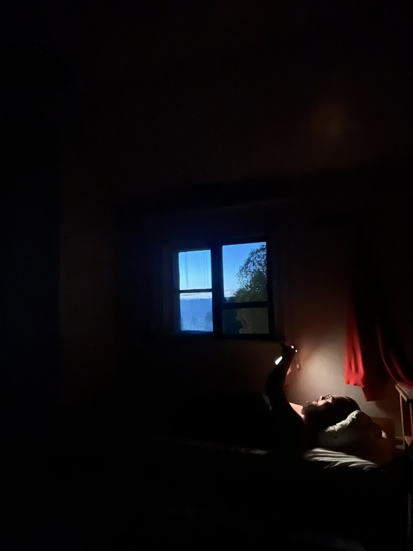 Bilden visar ett mörkt rum med ett litet fönster där blått skymningsljus strömmar in. Kontrasten mellan mörkret inne och ljuset ute skapar en stillsam, nästan drömlik stämning som kan väcka känslor av lugn, ensamhet eller eftertanke