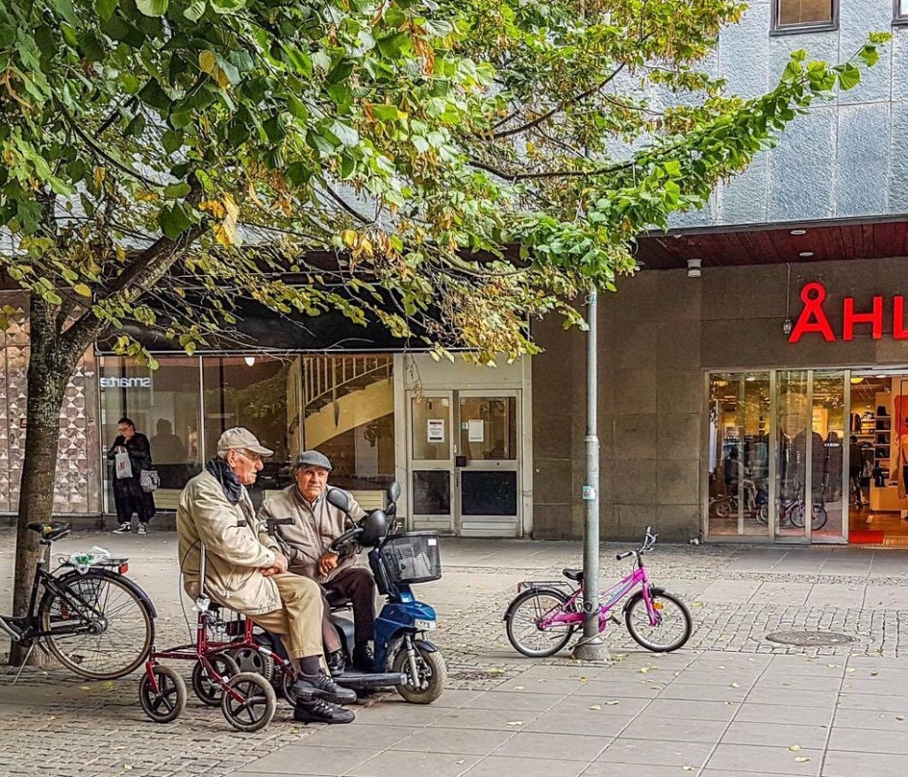 Två män med rullatorer på ett torg.
