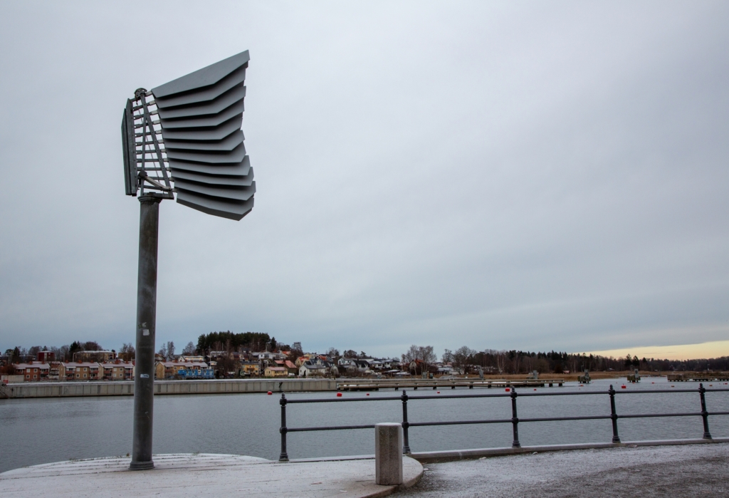 Skulptur i grått stål som veckar sig mot en ljusgrå himmel och vatten.