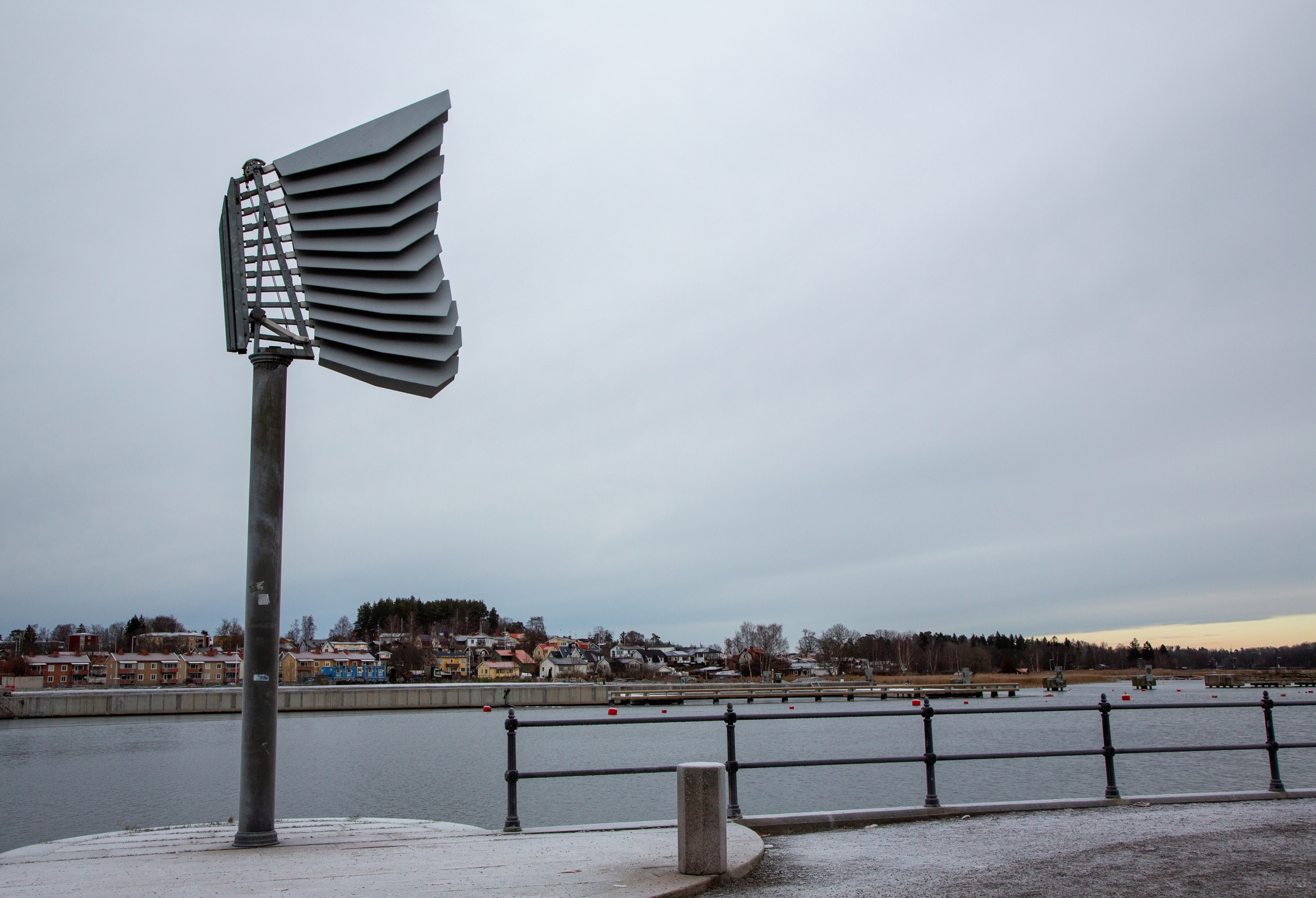 Skulptur i grått stål som veckar sig mot en ljusgrå himmel och vatten.