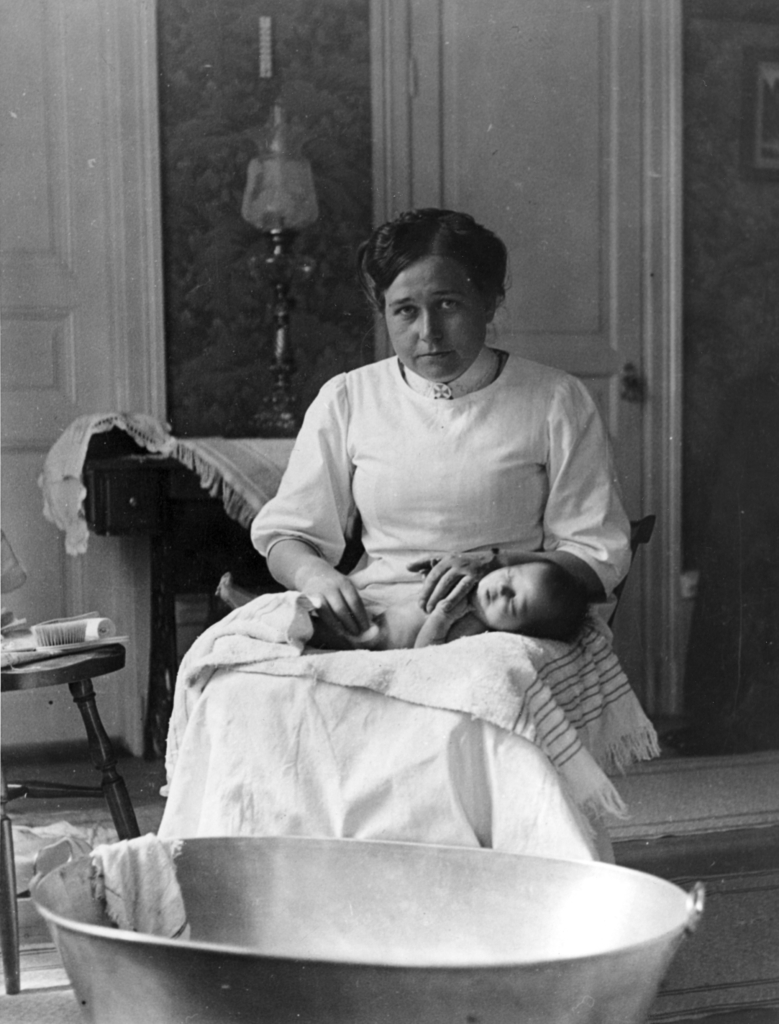 Historisk bild på en barnmorska med en nyfödd över knät i hemmamiljö.