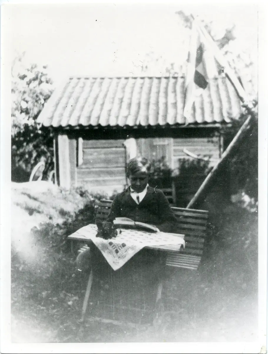 Svartvitt bleknat foto. En pojke sitter utomhus vid ett bor och läser en bok.