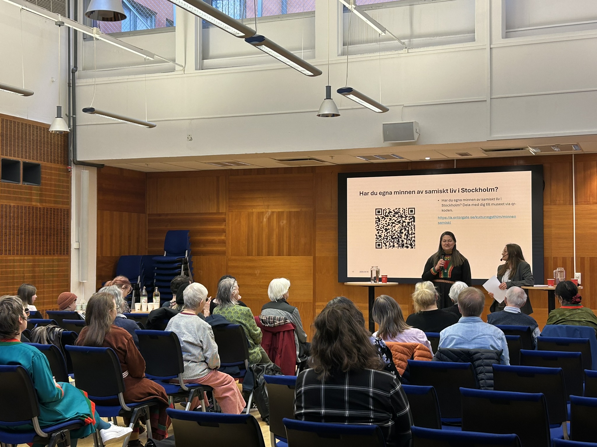 Föreläsningssal. Två kvinnor står längst fram och pratar. En bär samisk kolt. Publik tittar på.