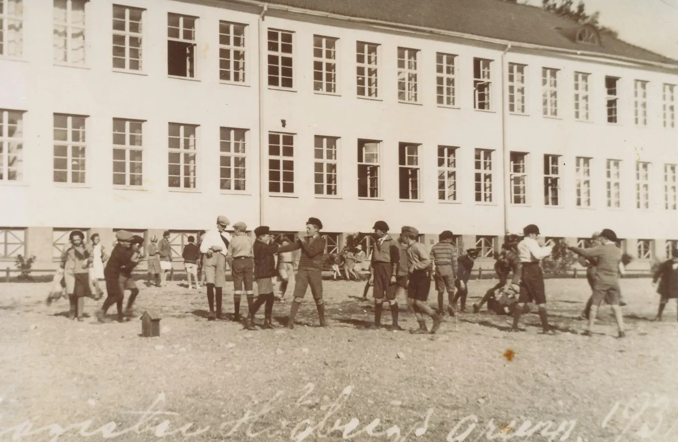 Barn framför skolans ljusa stora fasad. Flera pojkar verkar slåss. De bär keps, korta byxor och långa strumpor.