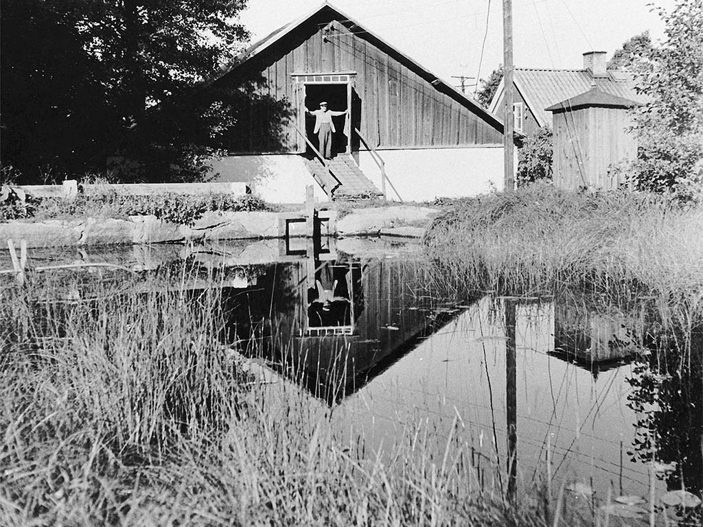 Svartvitt fotografi med en byggnad framför vatten. En man står i dörröppningen.