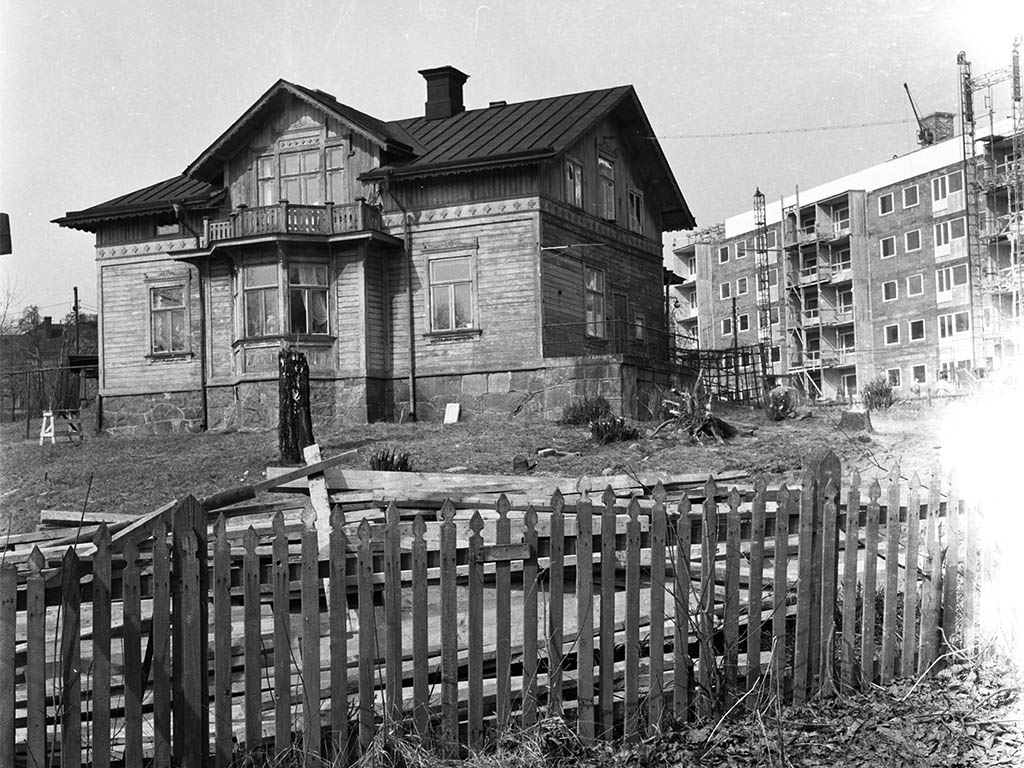 Svartvit bild med ett staket och ett trähus. och ett höghusbygge bakom