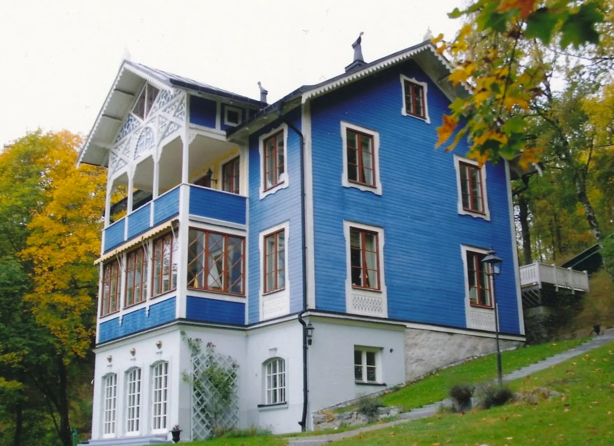 Ett blått trähus med två våningar och souterrain-våning. Stor balkong och veranda.