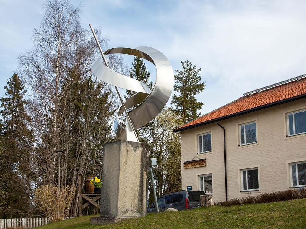 Blänkande abstrakt skulptur i stål framför ett gult hus.
