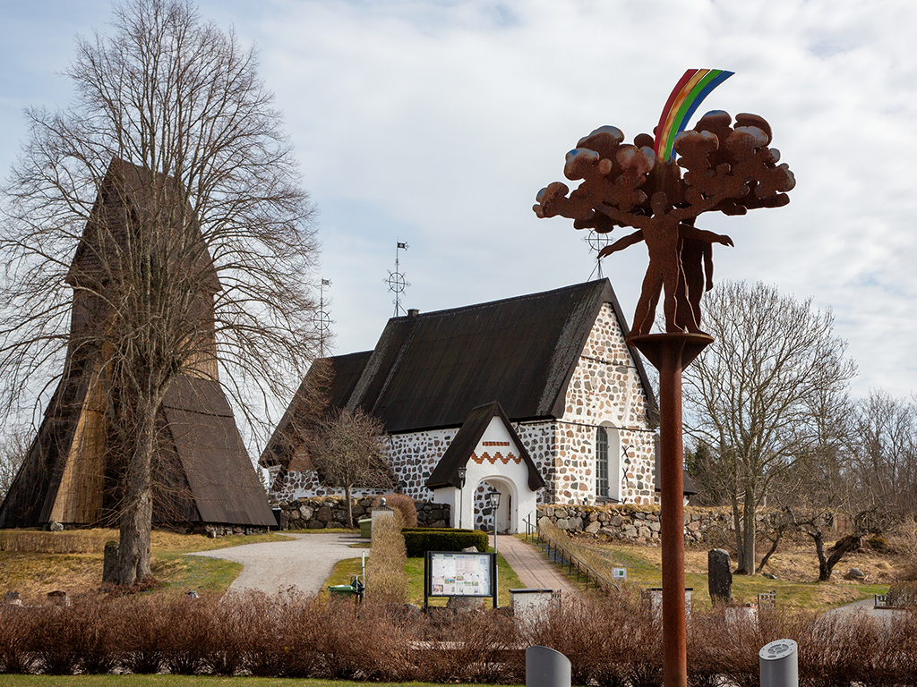 Skulptur i rostfärgad cortenstål med en människa som har en färgglad regnbåge och bubbliga former ovanför huvudet, med en gammal murad kyrka i bakom. .