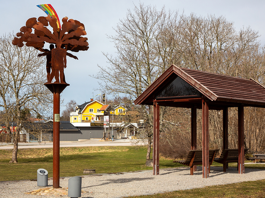 Hög skulptur i rostfärgat stål med en människa som har en färgglad regnbåge och bubbliga former ovanför huvudet, med en lite kur bredvid.