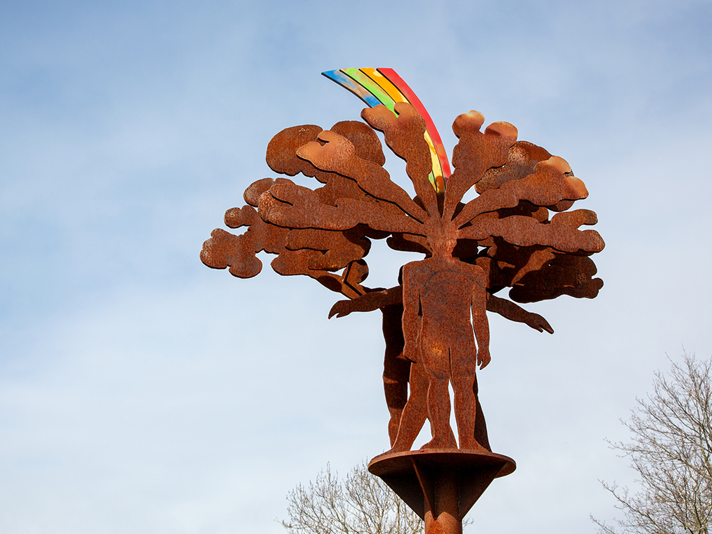 Skulptur i rostfärgat stål med en människa som har en färgglad regnbåge och bubbliga former ovanför huvudet och som syns mot en blå himmel.