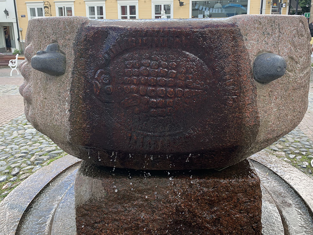Rund fontän på en sockel med rinnande vatten och med fisk som är inristad.