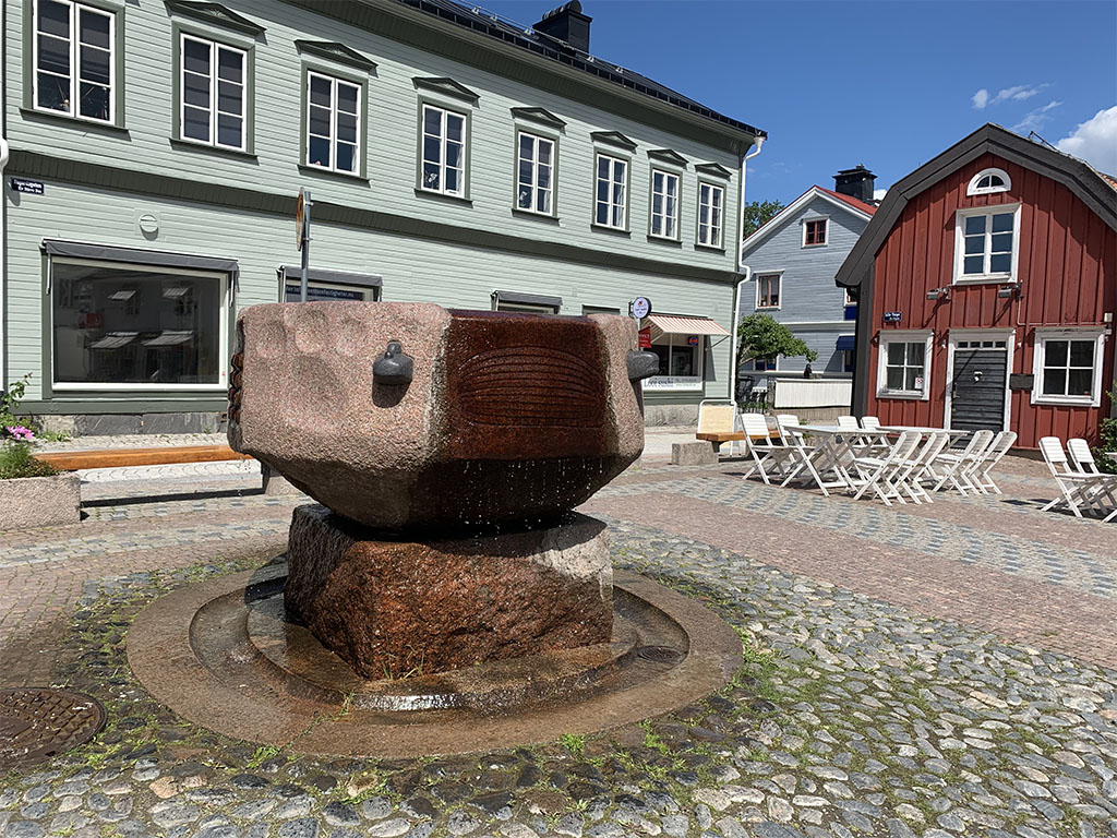 Fontän av röd sten på en sockel på ett torg med byggnader.