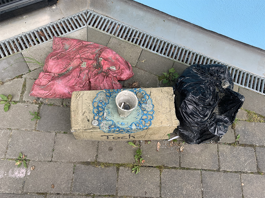 Tredelat konstverk utomhus med brun kartong, svart sopsäck och en skrynklig kudde. Sett ovanifrån.