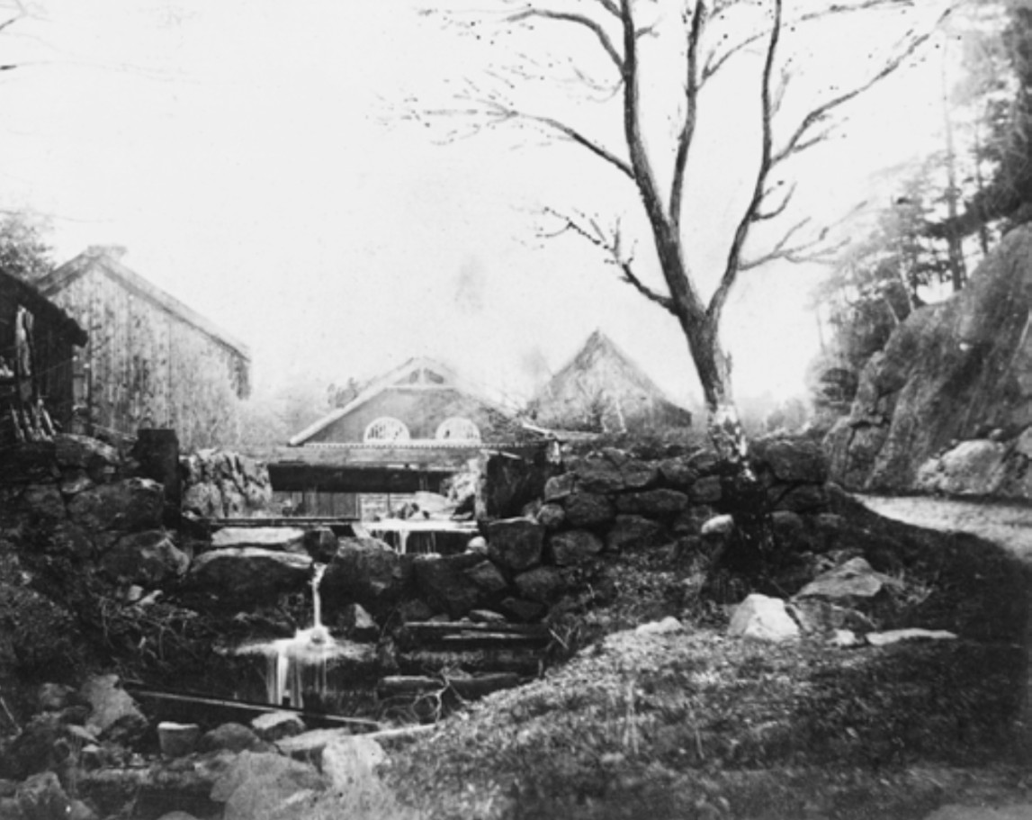 Svartvitt foto av kvarnmiljön med flera byggnader och strömmande vatten.