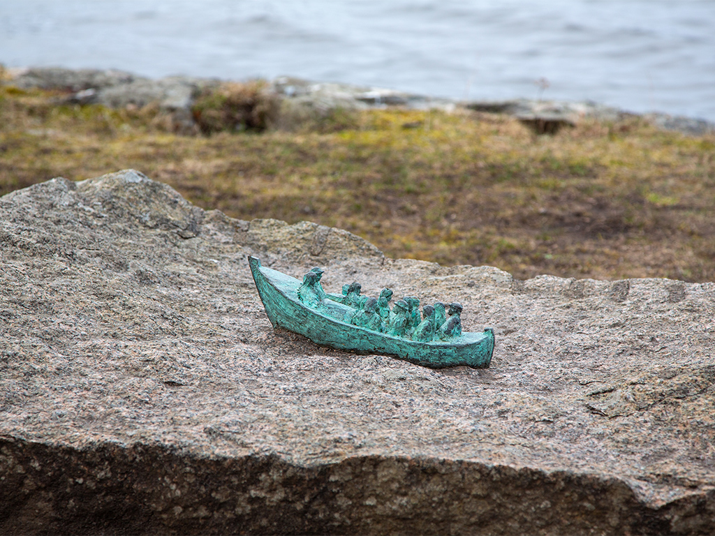 Skulptur av en liten grönaktig båt med människor på en stor sten intill vatten.