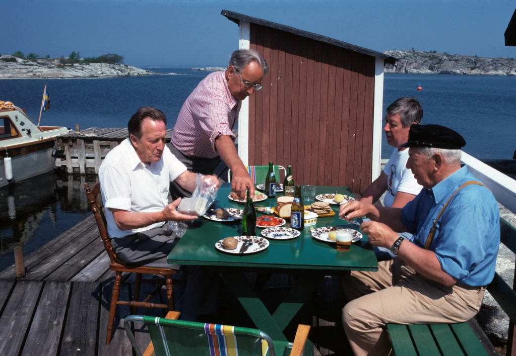 Fyra män vid ett uppdukat bord i skärgården. På bordet står tallrikar med fisk och potatis och flaskor med vad som skulle kunna vara öl.