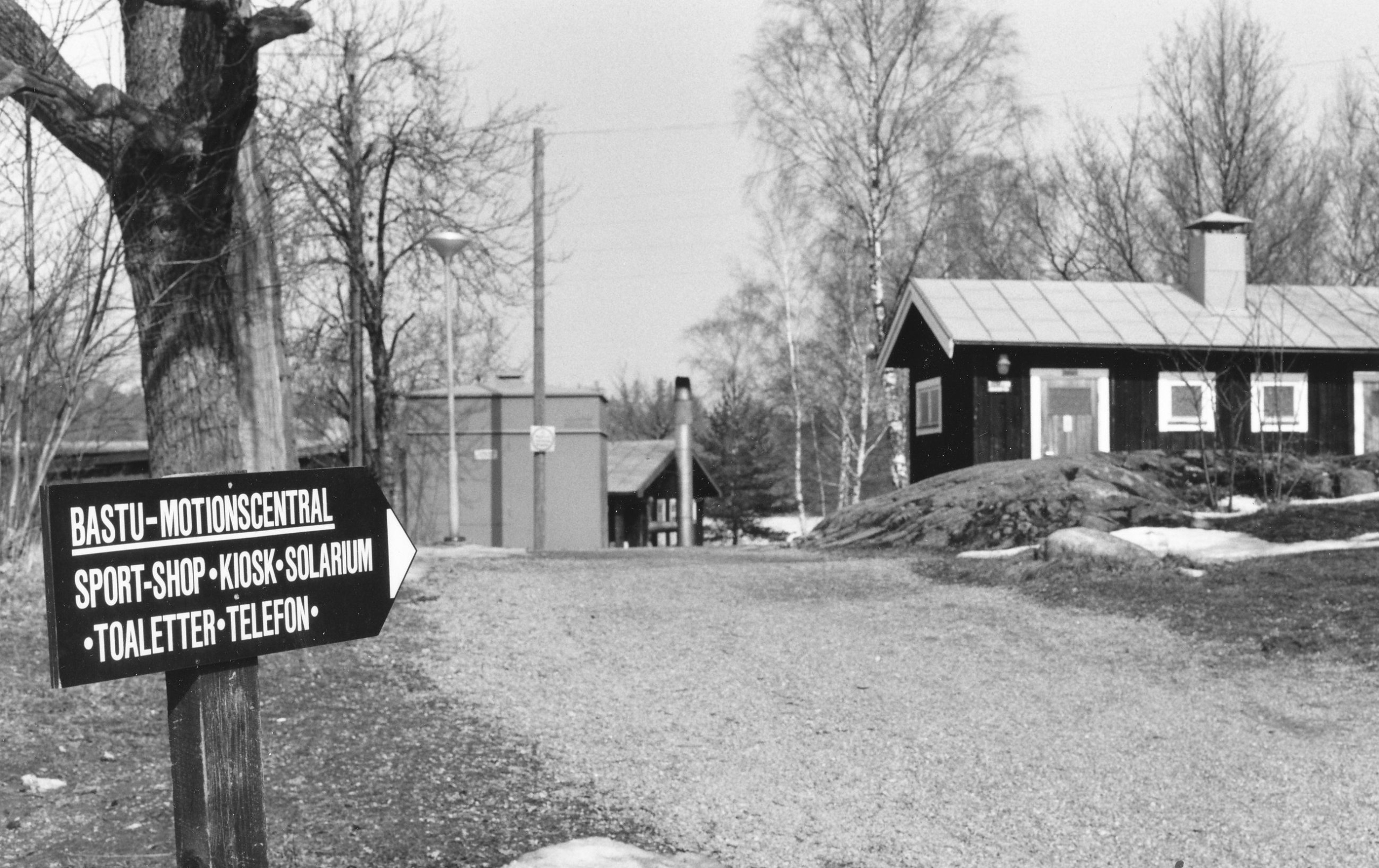 Närmast i bild är finns en skylt som visar vägen till bastun, shopen och toaletter. En grusväg leder bort, förbi ett hus.